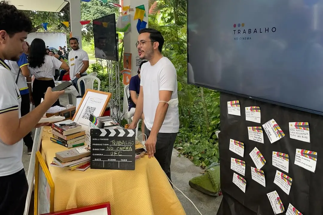 Um jovem de camiseta branca e óculos sorri ao lado de uma mesa com livros e uma claquete do projeto 'Trabalho no Cinema' da UFC. Ao fundo, uma tela exibe a logo do evento e um painel com cartões coloridos, compondo um cenário educativo e cultural em um ambiente aberto e arborizado.