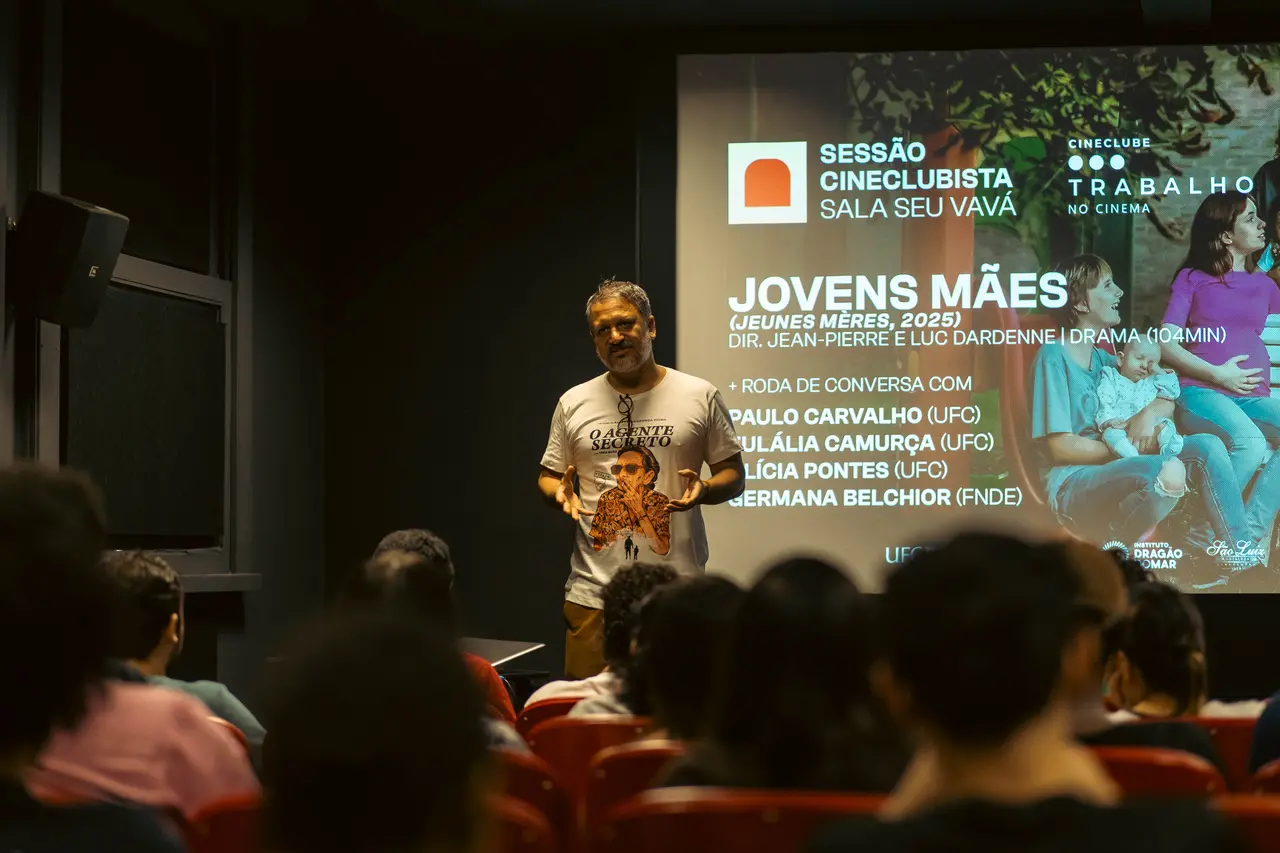 O professor Paulo Carvalho está de pé, de camiseta branca com a estampa do filme 'O Agente Secreto', e fala para uma plateia em um auditório escuro durante a exibição de 'Jovens Mães'. A projeção ao fundo detalha a sessão cineclubista e os participantes da roda de conversa, destacando um momento de debate e mediação cultural.