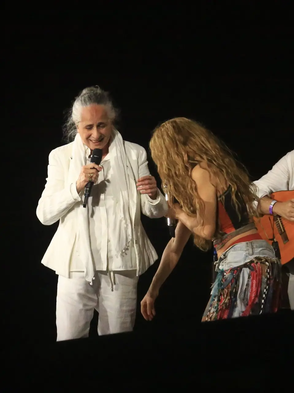 Mulher de cabelos grisalhos e roupa branca com microfone cantando ao lado de mulher loira de blusa preta curta e calça jeans.