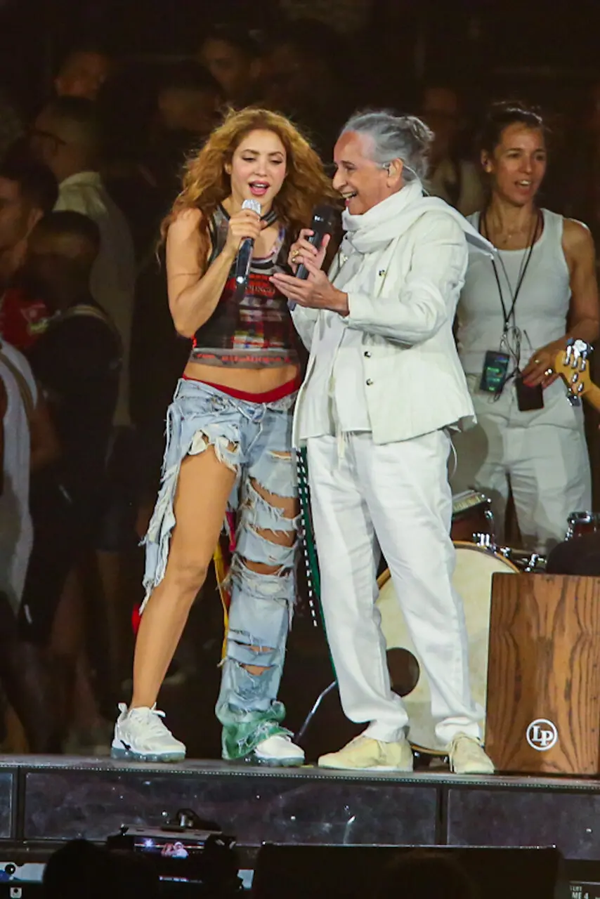 Mulher de cabelos grisalhos e roupa branca com microfone cantando ao lado de mulher loira de blusa preta curta e calça jeans.