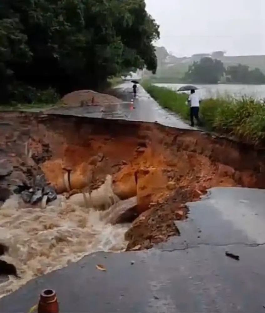 Na imagem, fotografia de um colapso em uma estrada asfáltica. Um enorme buraco corta a via de um lado ao outro, revelando o solo de terra alaranjada e tubulações de concreto rompidas. Uma enxurrada de água marrom flui intensamente pela base da cratera. Ao fundo, pedestres seguram guarda-chuvas perto de uma área de vegetação e um corpo d'água, indicando tempo chuvoso.