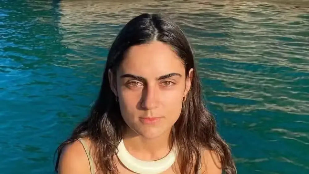 Mulher com cabelo castanho escuro, expressão séria, ao ar livre próximo a uma piscina ou corpo de água, fundo com tons de azul e verde