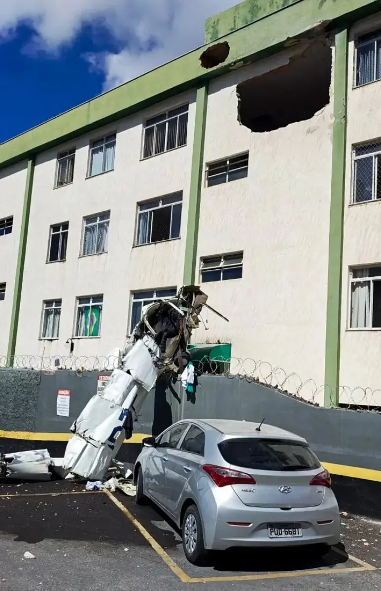 Destroços de avião que caiu em BH preso ao muro de um prédio, com uma parte pendurada e outra caída no chão; um carro está estacionado logo abaixo e há buracos na fachada do edifício.