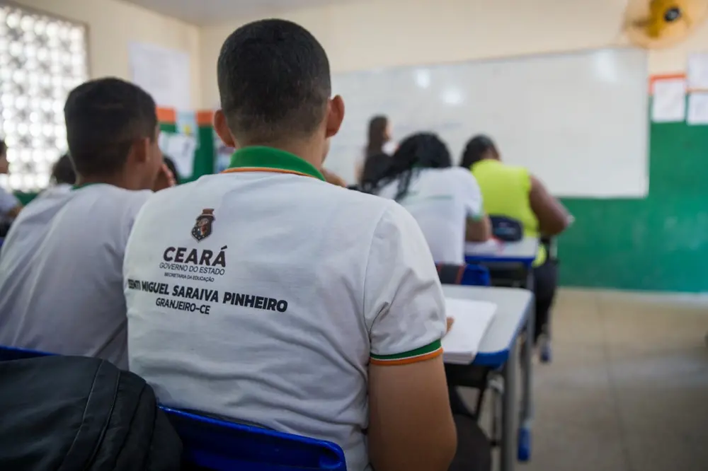 imagem de alunos em uma sala de aula com foco nas costas de um aluno com fardamento escolar.