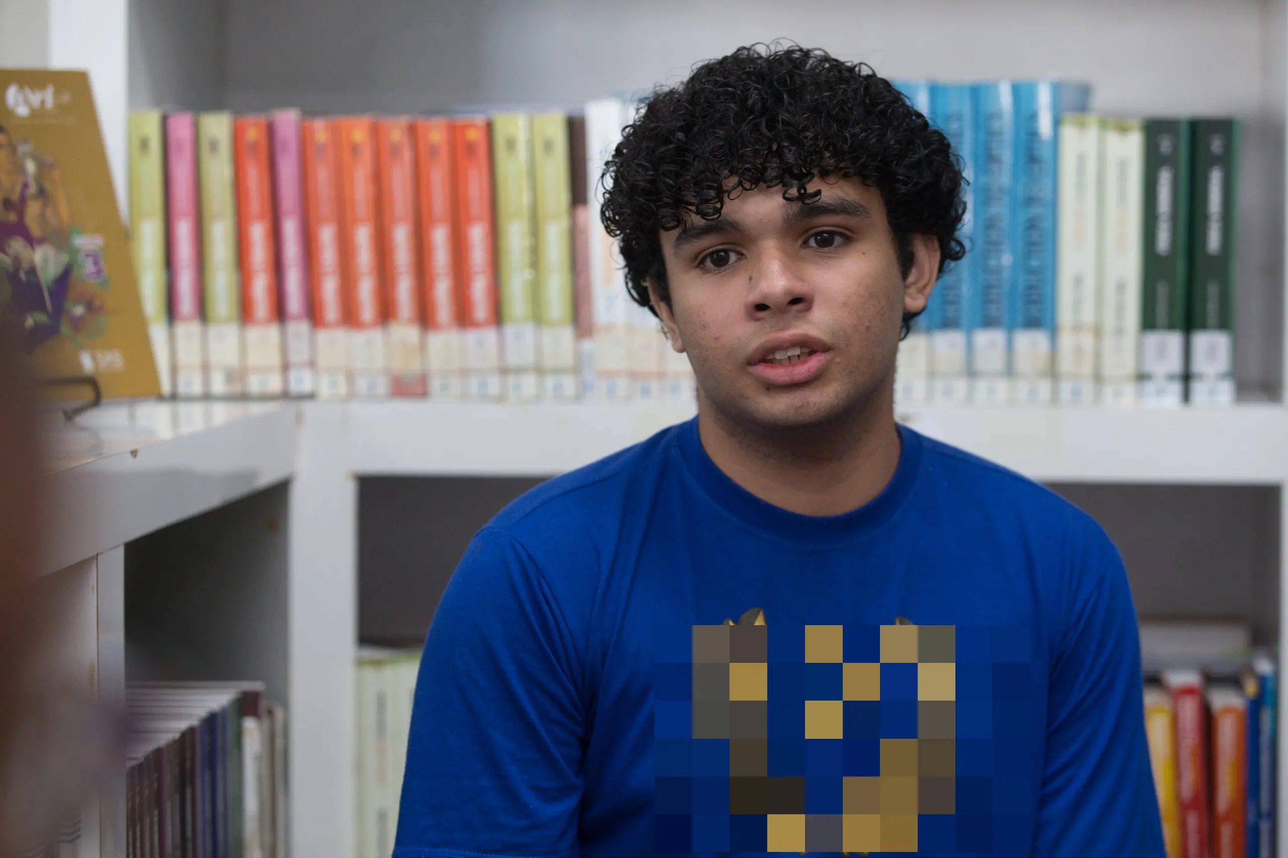 Um jovem de cabelos cacheados e camiseta azul com um brasão dourado está sentado em uma biblioteca, olhando para a câmera com uma expressão atenta. Ele está posicionado em primeiro plano, ladeado por prateleiras brancas repletas de livros coloridos que preenchem todo o fundo da imagem. A iluminação é natural e clara, focando no rosto do estudante e no ambiente de estudos ao seu redor.