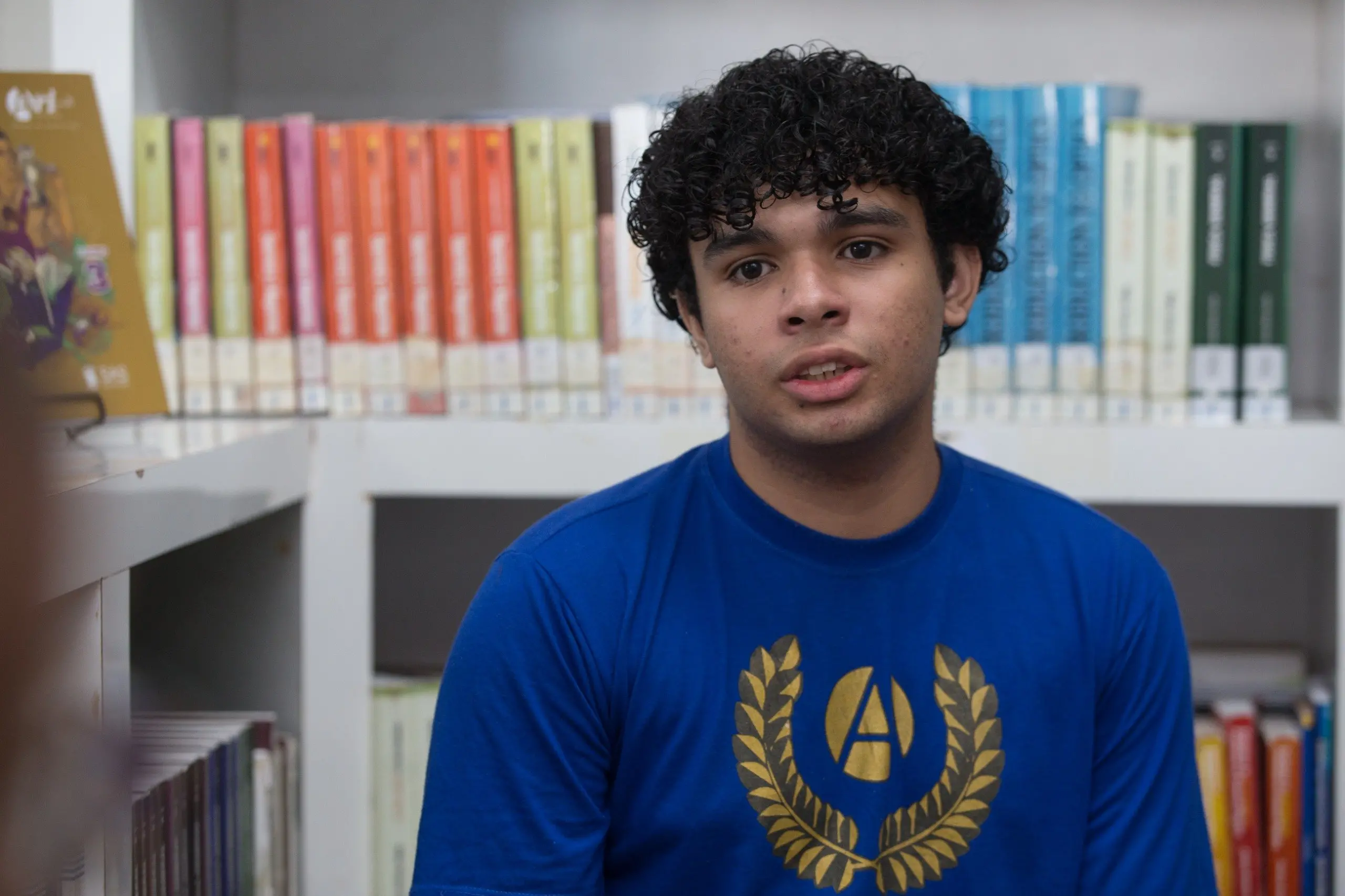 Um jovem de cabelos cacheados e camiseta azul com um brasão dourado está sentado em uma biblioteca, olhando para a câmera com uma expressão atenta. Ele está posicionado em primeiro plano, ladeado por prateleiras brancas repletas de livros coloridos que preenchem todo o fundo da imagem. A iluminação é natural e clara, focando no rosto do estudante e no ambiente de estudos ao seu redor.