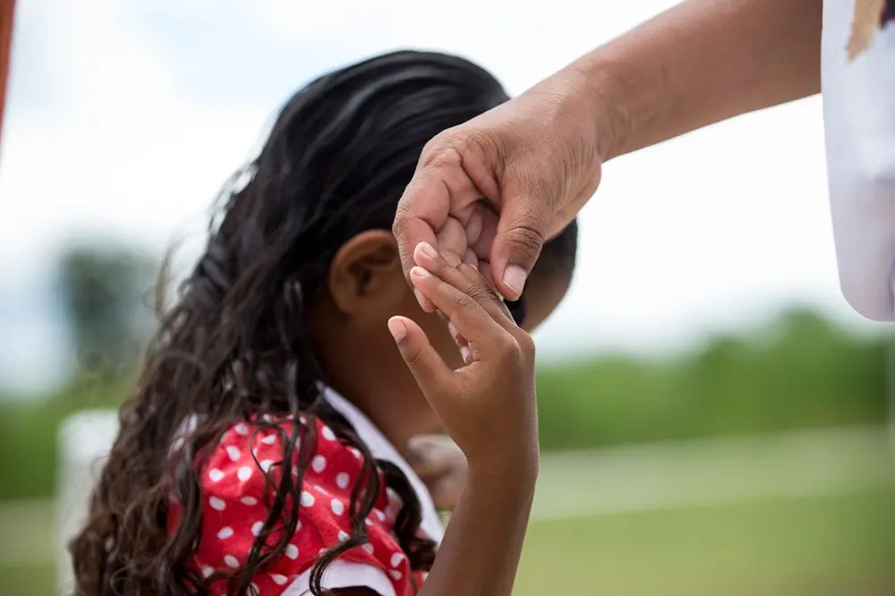 Na imagem, close-up emocionante que captura o toque entre as mãos de um adulto e de uma criança. A mão do adulto, posicionada na parte superior, segura delicadamente os dedos da criança. A criança, vista de costas e levemente desfocada, tem cabelos longos e escuros e veste o traje tradicional de devoção à Beata Benigna: um vestido vermelho com estampa de bolinhas brancas. O fundo é composto por tons de verde e um céu claro, criando uma atmosfera suave e de cuidado.