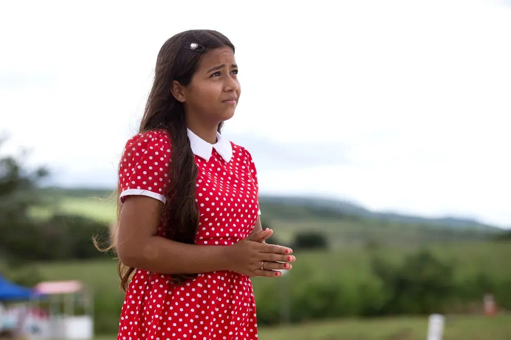 Na imagem, plano médio de uma menina adolescente com as mãos postas em sinal de oração. Ela tem cabelos longos e castanhos, usa uma presilha delicada e olha para cima com uma expressão de devoção e esperança. A jovem veste o traje característico da Beata Benigna: um vestido vermelho com estampa de bolinhas brancas e gola branca. O fundo mostra uma paisagem natural verdejante sob um céu claro, de forma suavemente desfocada.