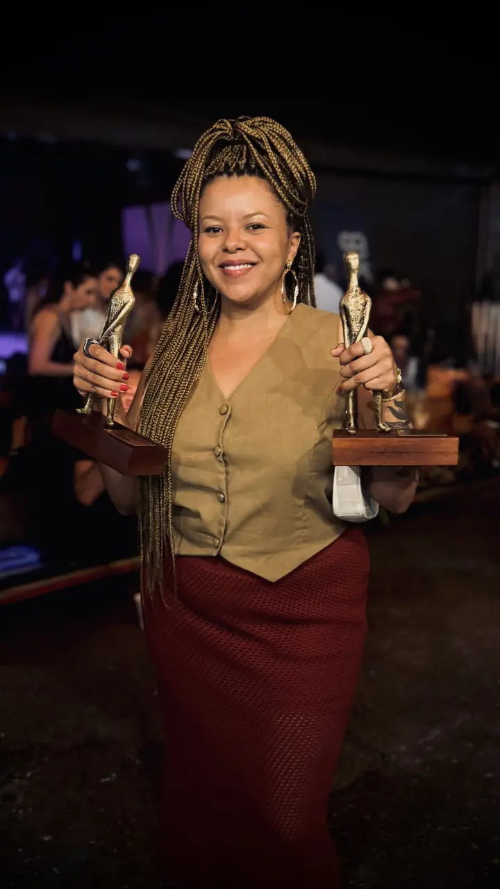 A cineasta Karol Maia sorri segurando dois troféus dourados, destacando suas longas tranças loiras e seu traje em tons de bege e vinho sob uma iluminação suave. A composição captura um momento de triunfo em um ambiente festivo e escuro, ressaltando o contraste entre o brilho das estatuetas e o fundo sutilmente desfocado.