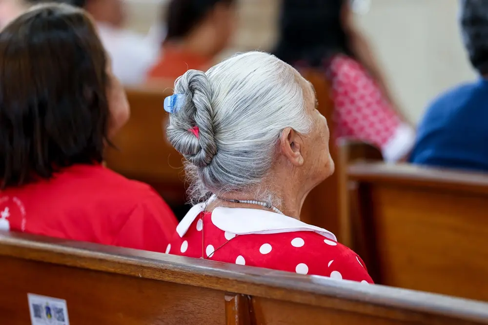 Na imagem, fotografia em close-up, tirada de costas e levemente de perfil, de uma senhora idosa com cabelos brancos presos em um coque elegante, fixado com um grampo azul. Ela está sentada em um banco de madeira de uma igreja, vestindo uma blusa vermelha com estampa de bolinhas brancas e gola boneca, em homenagem à vestimenta da Beata Benigna Cardoso. Ao fundo, de forma desfocada, aparecem outros fiéis sentados nos bancos, criando uma atmosfera de devoção e respeito. A iluminação é suave, destacando a textura do cabelo e o contraste das cores da roupa.
