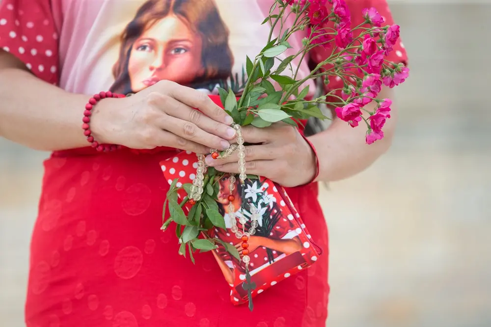 Na imagem, fotografia em plano detalhado das mãos de uma pessoa segurando objetos de devoção. Ela veste uma camiseta com a estampa do rosto da Beata Benigna Cardoso e uma saia vermelha. Entre as mãos, segura um pequeno buquê de flores rosas naturais, um terço de contas transparentes com um crucifixo escuro e uma pequena bolsa ou estojo vermelho com a imagem da Beata estampada. No pulso esquerdo, a pessoa usa uma pulseira de contas vermelhas. O fundo está desfocado em tons claros, mantendo o foco total nos símbolos de fé.