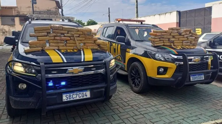Duas viaturas da Polícia Rodoviária Federal estacionadas em área urbana, com vários tabletes de entorpecentes empilhados sobre os capôs dos veículos.