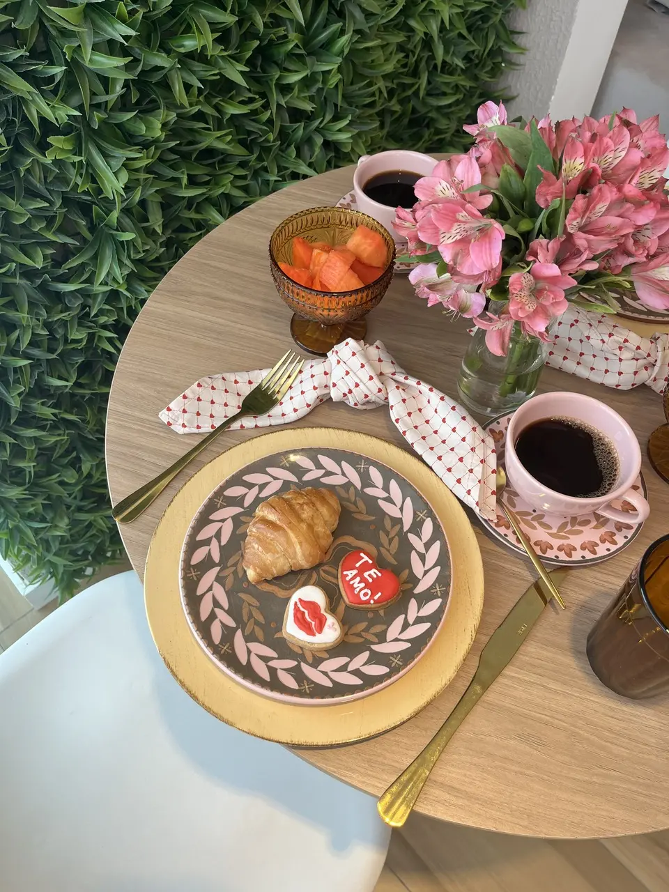 Mesa de café da manhã decorada com flores rosas, xícaras de café, croissant, frutas e biscoitos em formato de coração com a frase “Te amo”, sobre pratos estampados e talheres dourados.