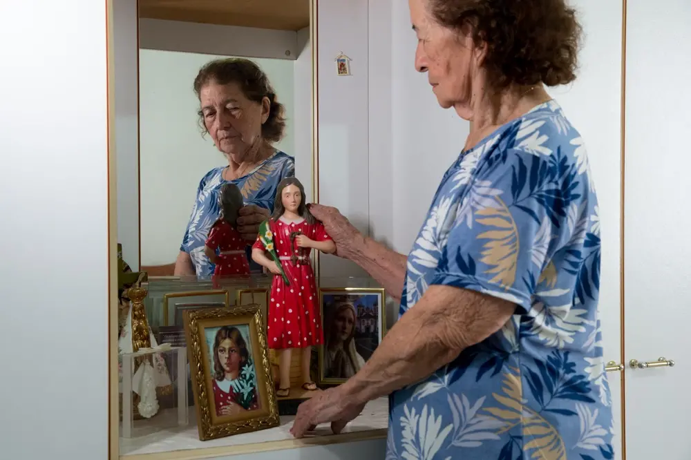 Na imagem, uma mulher idosa de cabelos castanhos e curtos, vestindo uma blusa azul com estampas de folhagens brancas e bege, está de pé em frente a uma penteadeira ou altar doméstico. Ela é vista de perfil e seu reflexo aparece no espelho à sua frente. Com a mão direita, ela toca suavemente a cabeça de uma pequena estátua da Beata Benigna, que veste um vestido vermelho com bolas brancas. Sobre a bancada, além da estátua, há um porta-retrato com a ilustração da Beata, uma imagem de Nossa Senhora de Fátima e outros objetos religiosos. O móvel é branco e a iluminação é suave e natural.