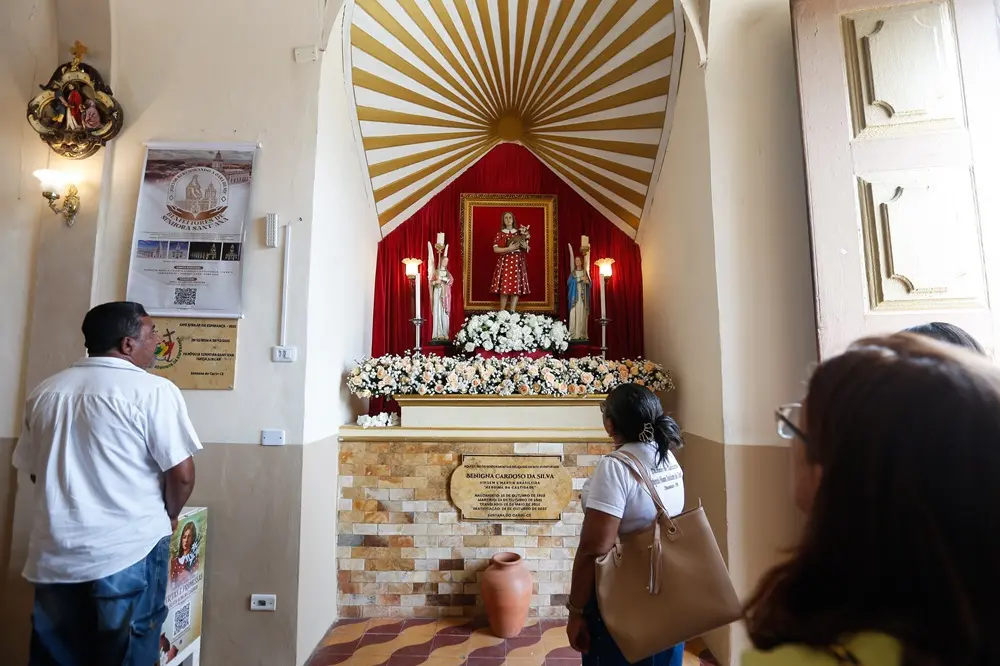 Na imagem, fotografia do interior de uma igreja mostrando o nicho dedicado à Beata Benigna Cardoso da Silva. No centro de um altar elevado, decorado com muitas flores brancas e rosas, destaca-se uma estátua da Beata vestindo seu icônico vestido vermelho com bolas brancas. Ela está emoldurada por um fundo de tecido vermelho vibrante e um arco superior com raios dourados e brancos. Abaixo da imagem, na parede de pedras, há uma placa comemorativa com informações sobre sua vida e martírio. À esquerda, um homem de camisa branca observa o altar, e à direita, uma mulher com uma bolsa bege e parte do rosto de outra fiel aparecem em primeiro plano, contemplando o local. Na parede lateral esquerda, há cartazes informativos e uma luminária clássica.