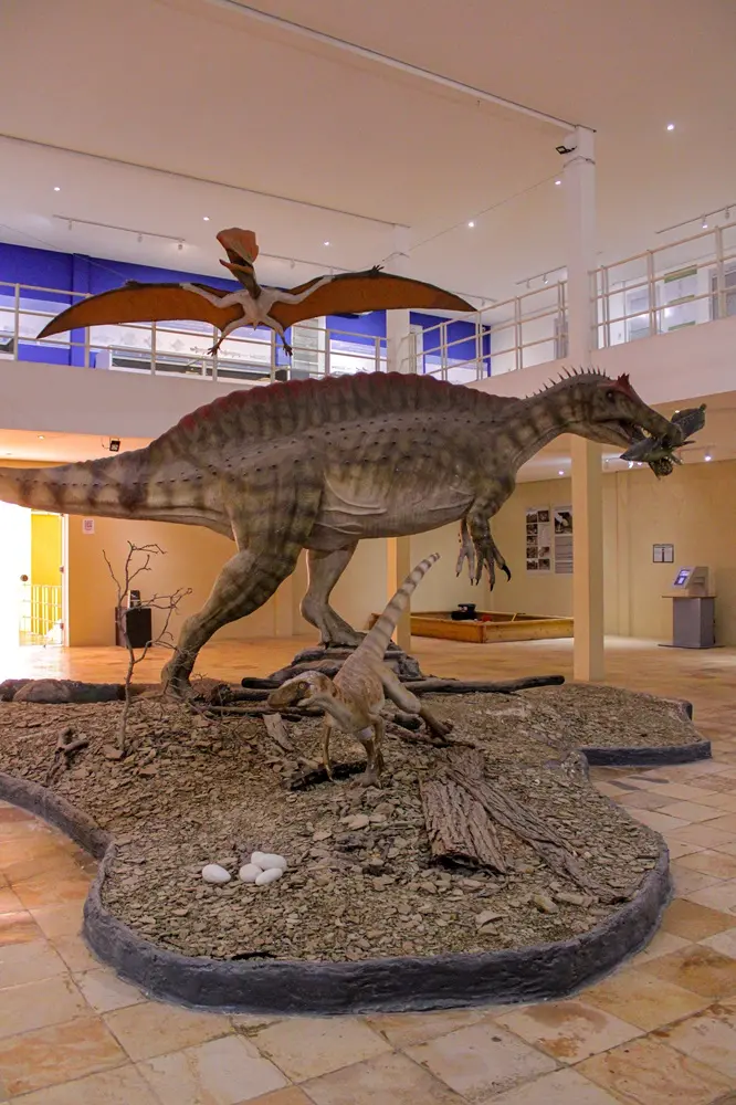 Na imagem, fotografia do interior de um museu com uma exposição de réplicas de dinossauros em tamanho real. No centro, sobre uma base de cascalho que simula o solo natural, destaca-se um grande espinossaurídeo com uma crista avermelhada nas costas, segurando um peixe na boca. À sua frente, há um dinossauro menor em posição de corrida e, no chão, alguns ovos brancos. Suspenso acima deles, um pterossauro de asas abertas parece sobrevoar a cena. O ambiente do museu possui paredes claras, mezanino com guarda-corpo branco e iluminação por focos de luz no teto.