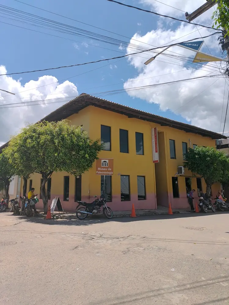Na imagem, fotografia da fachada externa do Museu de Paleontologia Plácido Cidade Nuvens, em Santana do Cariri. O prédio de dois pavimentos tem paredes amarelas com uma faixa rosa na base e telhado de barro marrom. Na esquina, há uma placa marrom com o desenho de um templo clássico e os dizeres 