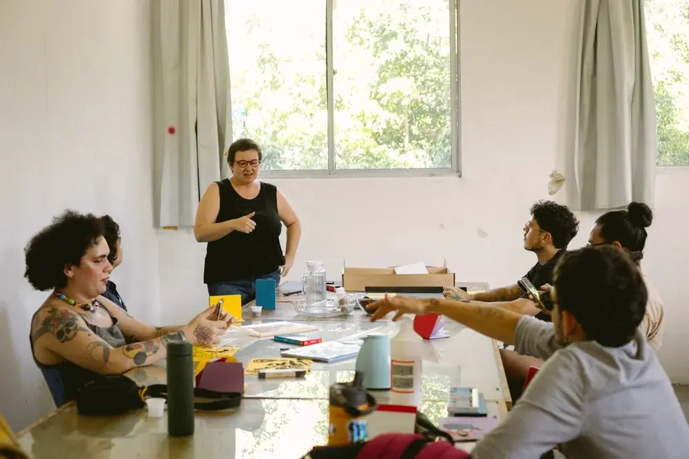 Imagem de atividade do Laboratório de Artes Visuais do Porto Iracema das Artes, que mostra um grupo reunido em torno de uma mesa de trabalho com materiais artísticos. A cena ocorre em uma sala clara e minimalista, com luz natural vindo de uma janela ao fundo, capturando um momento de aprendizado coletivo e troca criativa.