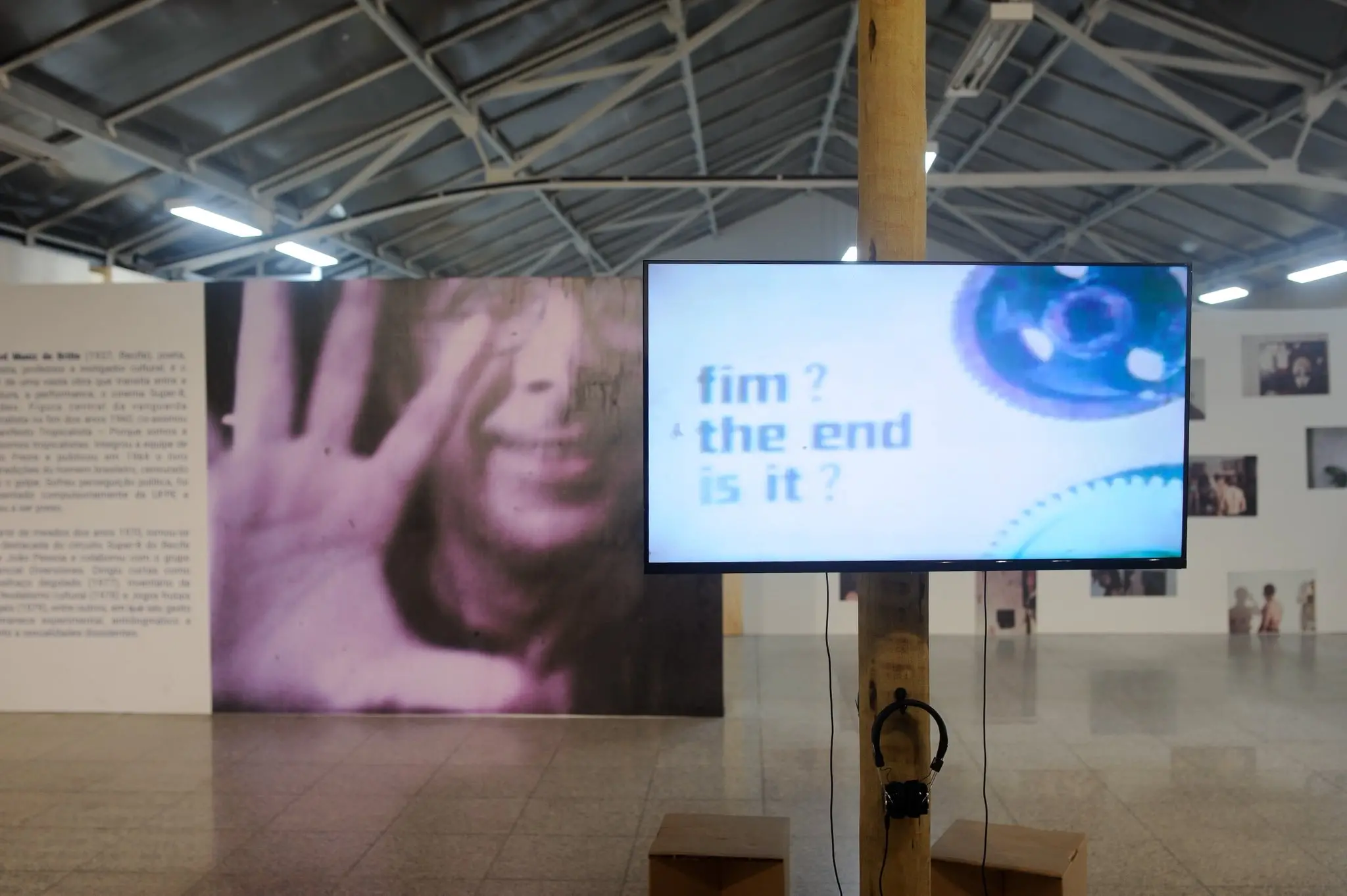 Registro da exposição Arquivo em Fluxo, no Banco do Nordeste Cultural Fortaleza, mostra uma tela de televisão montada em um pilar de madeira exibe as palavras 'fim? the end is it?' em uma galeria de arte. Ao fundo, um grande painel apresenta o rosto de um homem com a mão aberta à frente, cercado por textos biográficos e outras fotografias da exposição.