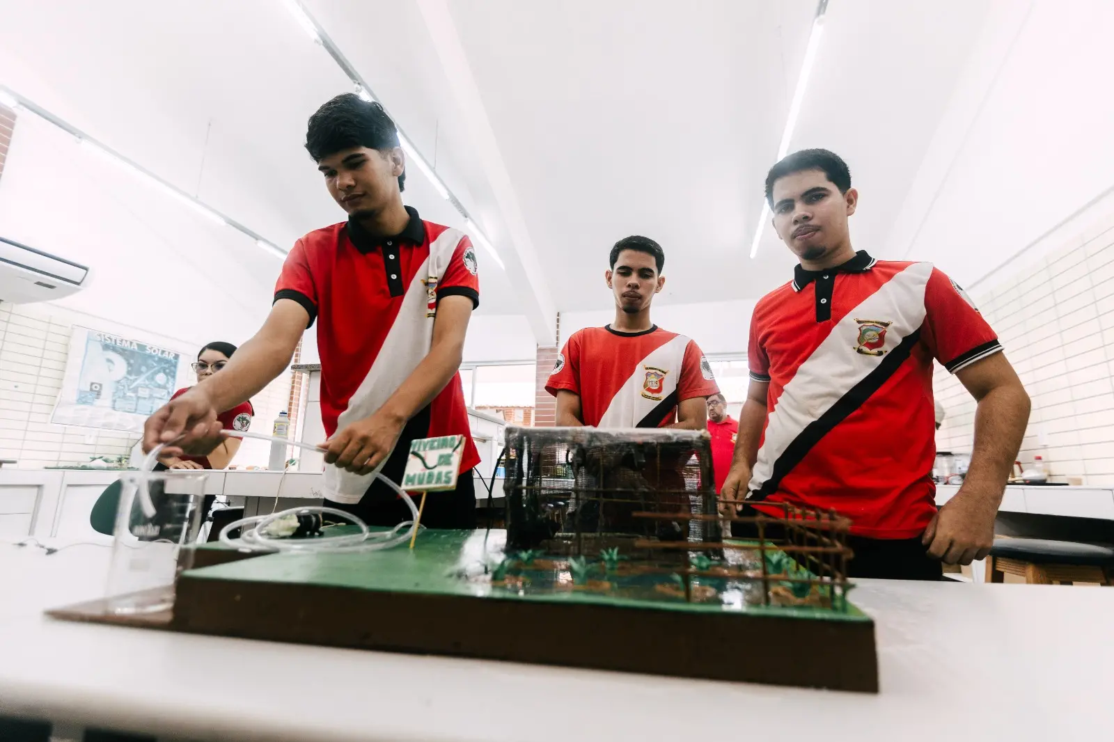 imagem de três jovens estudantes mexendo em um circuito eletrônico em cima de uma mesa. O circuito está em primeiro plano e os estudantes em segundo.
