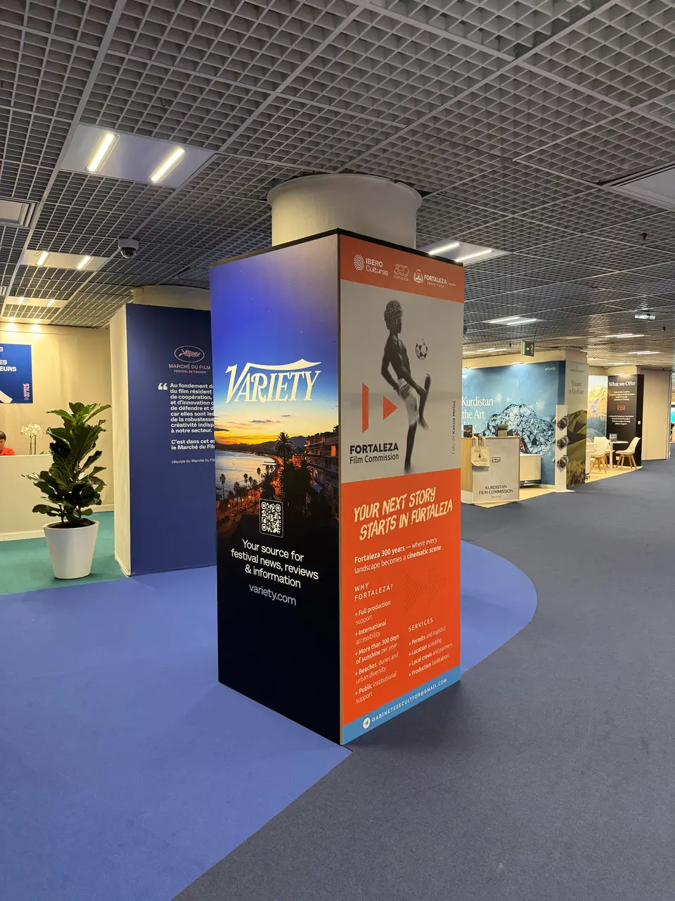 Um totem publicitário de quatro faces destaca as marcas da revista Variety e da Fortaleza Film Commission nos corredores de um pavilhão com carpete azul. Uma das faces exibe o logotipo da Variety sobre uma foto de um pôr do sol litorâneo, enquanto a face ao lado, em tons de branco e laranja, traz a imagem de um jovem jogando futebol e informações institucionais sobre Fortaleza.
