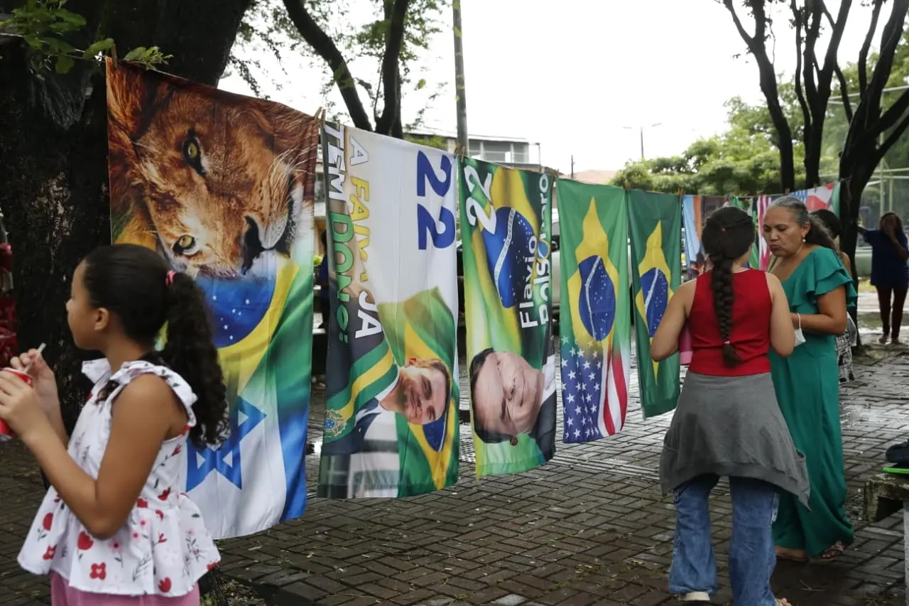Bandeiras com símbolos políticos e nacionais são exibidas em espaço público enquanto pessoas circulam pelo local durante evento ao ar livre.
