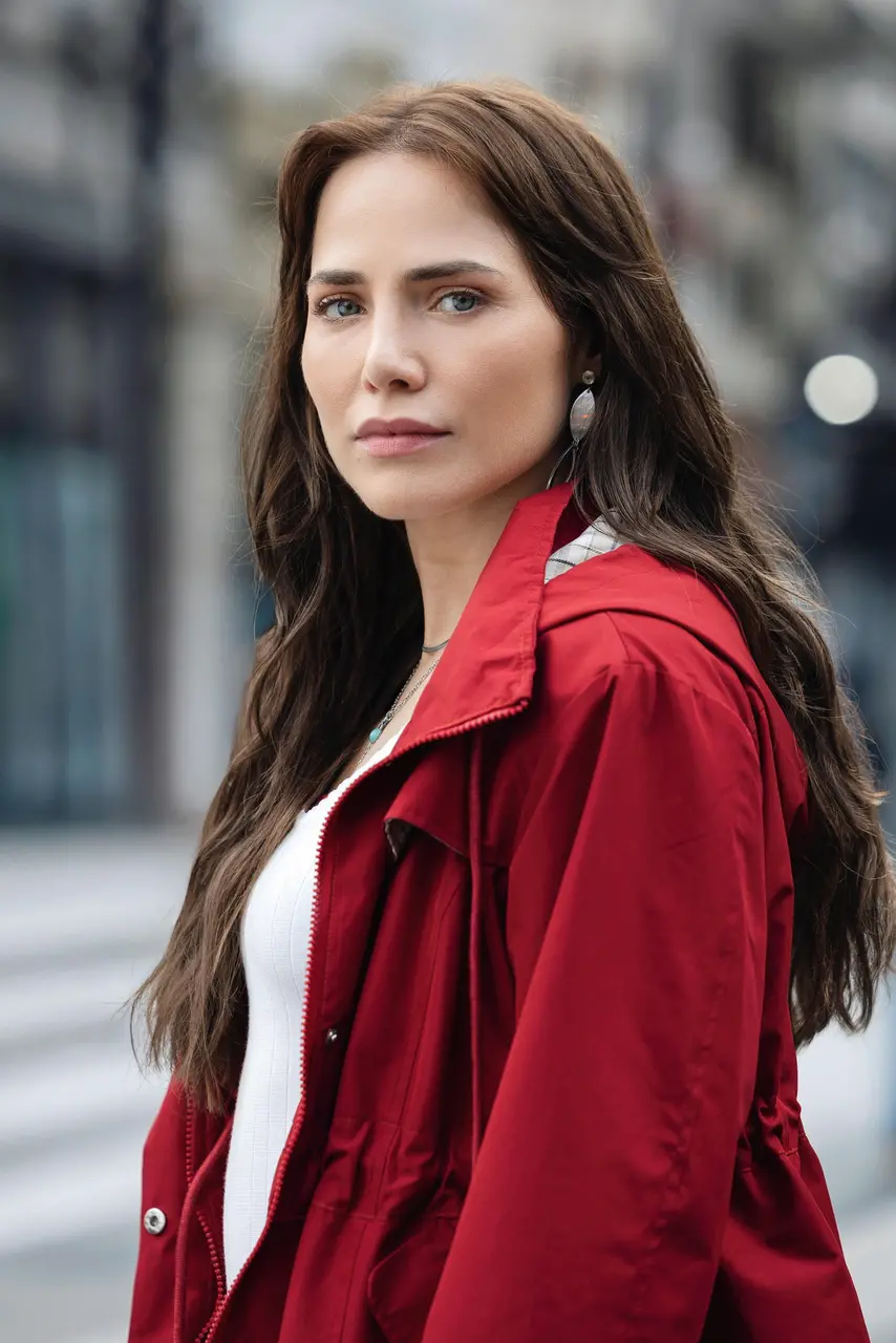 Pessoa com cabelo castanho longo posa em uma rua urbana desfocada ao fundo, vestindo casaco vermelho aberto sobre blusa clara. A pessoa está posicionada em primeiro plano, com leve inclinação do corpo para o lado, e usa brinco metálico visível. A iluminação é natural e suave, com tons neutros no cenário contrastando com a cor vibrante da roupa.