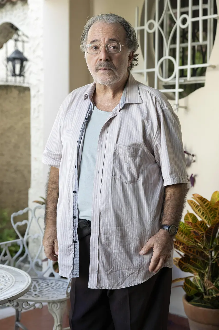 Pessoa em primeiro plano está em uma varanda ou área externa de residência, com parede clara e janela com grades decorativas ao fundo. A pessoa usa óculos, camisa clara aberta sobre uma blusa em tom neutro e calça escura. Há um relógio no pulso. Ao redor, aparecem móveis de metal pintados de branco e plantas em vasos. A iluminação é natural, com tons suaves e ambiente bem iluminado, destacando os elementos do espaço doméstico.