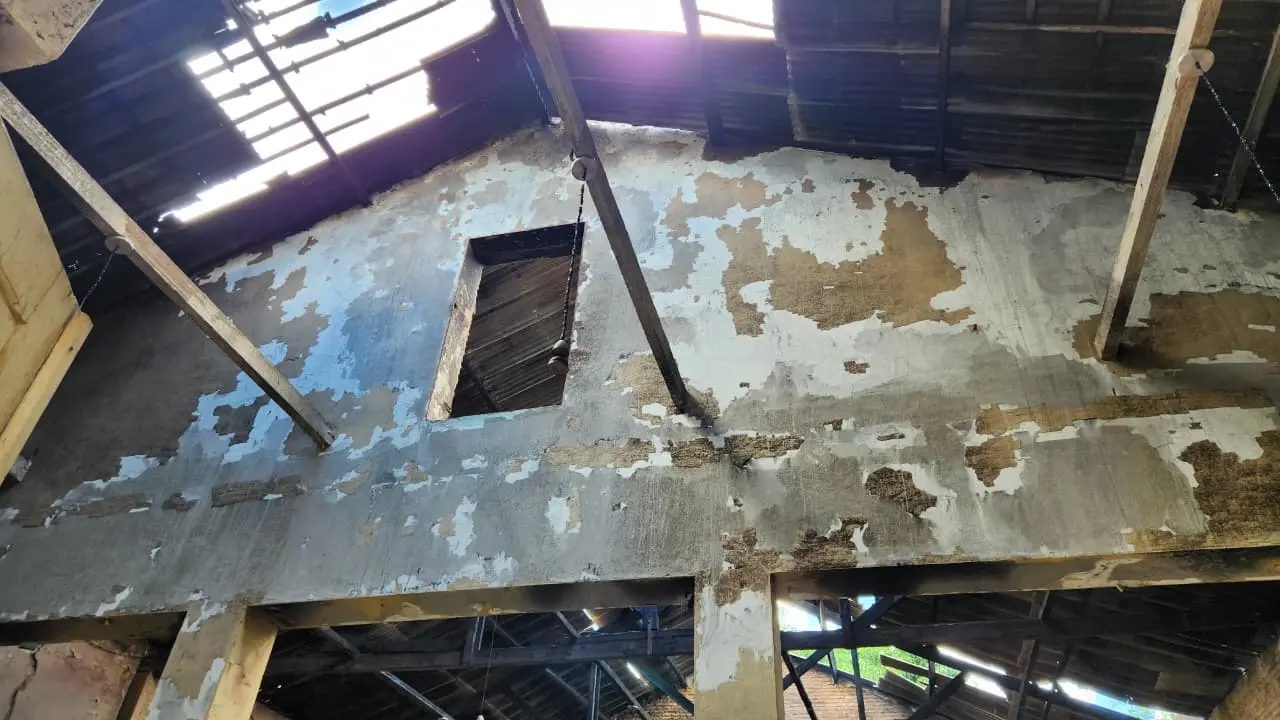 Interior de uma construção antiga em processo de reforma, com piso e paredes descascados e áreas de reboco e concreto expostos, com abertura no teto e vigas de madeira.