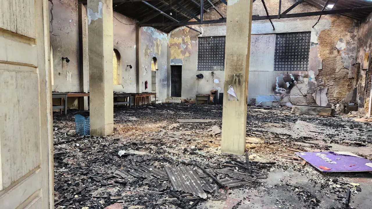 Interior de prédio antigo abandonado com paredes descascadas, tijolos aparentes e piso coberto por escombros e poeira, com pilares de concreto e salas vazias.