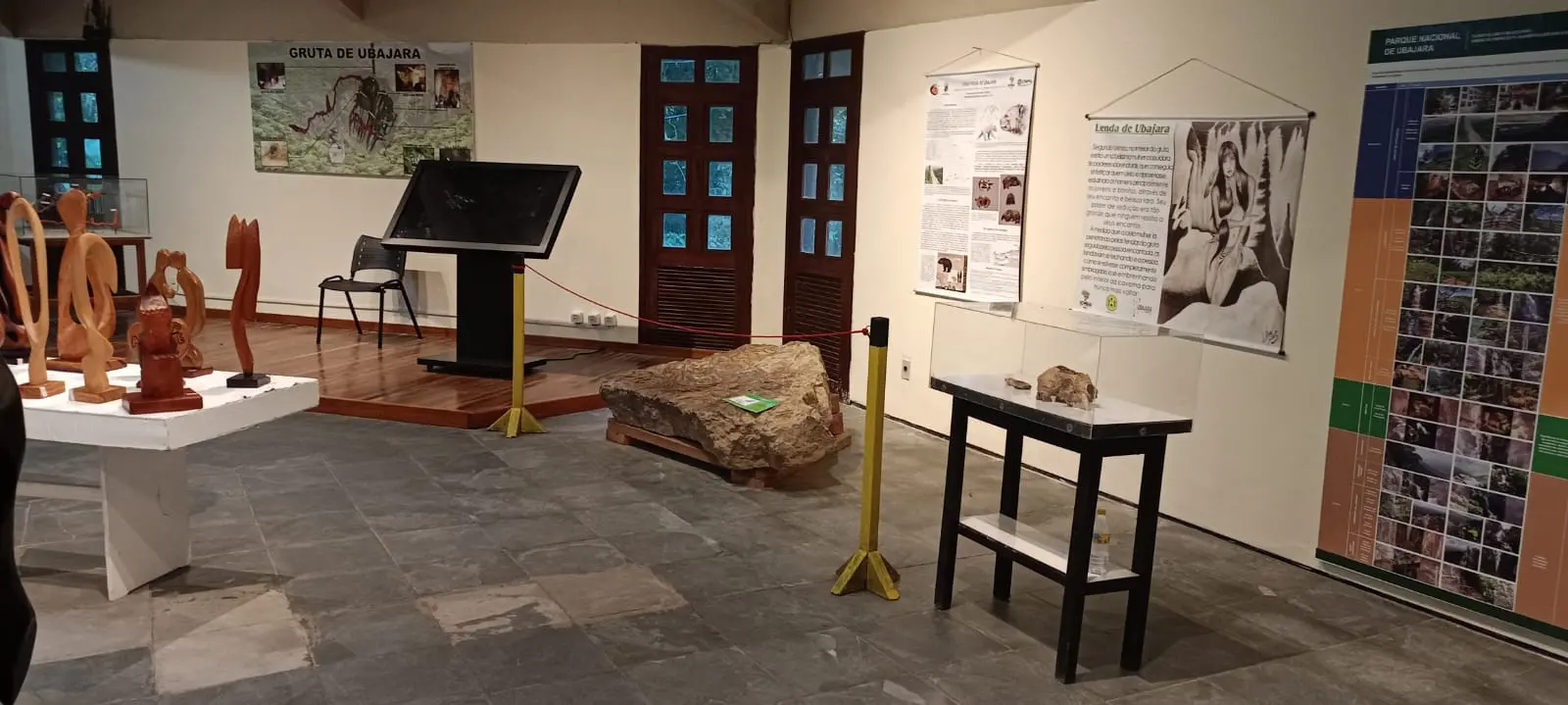 Fotografia em plano aberto de uma sala de exposição de museu com piso cinza e paredes brancas decoradas com painéis informativos sobre a Gruta de Ubajara. O espaço exibe esculturas de madeira em uma mesa à esquerda, uma grande rocha no centro isolada por pedestais amarelos e cordas vermelhas, e uma vitrine de vidro sobre uma mesa preta à direita.