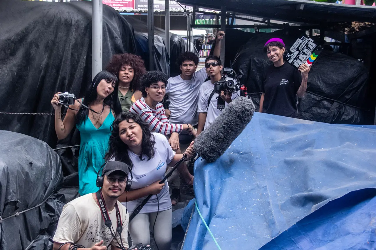 Uma jovem equipe de cinema posa sorridente e descontraída em um ambiente externo cercado por lonas pretas e azuis, segurando equipamentos como câmera fotográfica, microfone boom e uma claquete. O grupo diversificado de jovens demonstra entusiasmo nos bastidores da produção cinematográfica, combinando poses criativas em uma composição dinâmica e cheia de energia.