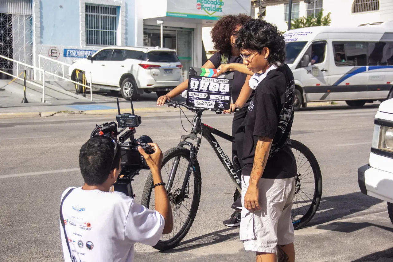 Integrantes de uma equipe de cinema gravam uma cena na rua, destacando um operador de câmera agachado em primeiro plano e uma jovem segurando a claquete à frente de uma bicicleta. O cenário urbano ao fundo traz carros estacionados, uma van em movimento e fachadas comerciais sob a luz do dia, capturando a dinâmica da produção audiovisual.
