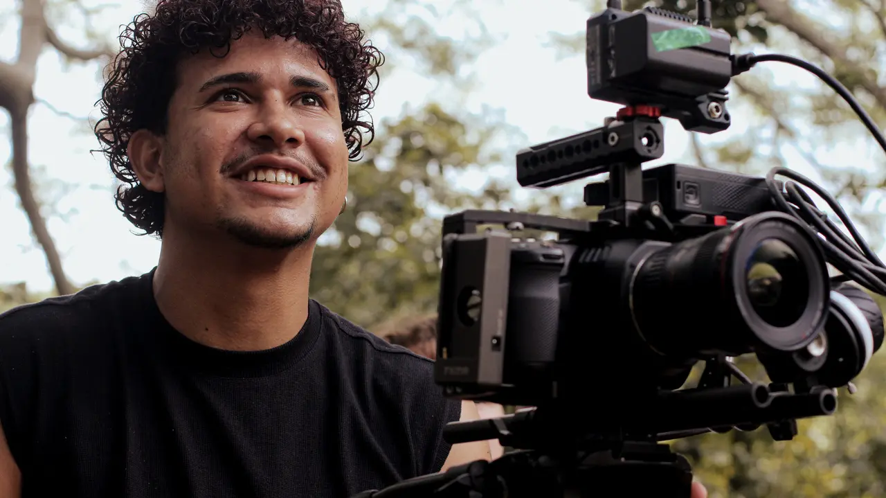 Um jovem de cabelos cacheados sorri com entusiasmo enquanto opera uma câmera de cinema profissional montada em um tripé nos bastidores de uma filmagem. O plano médio destaca a expressão de satisfação do cinegrafista em primeiro plano, contra um fundo verde e luminoso composto por árvores e folhagens.