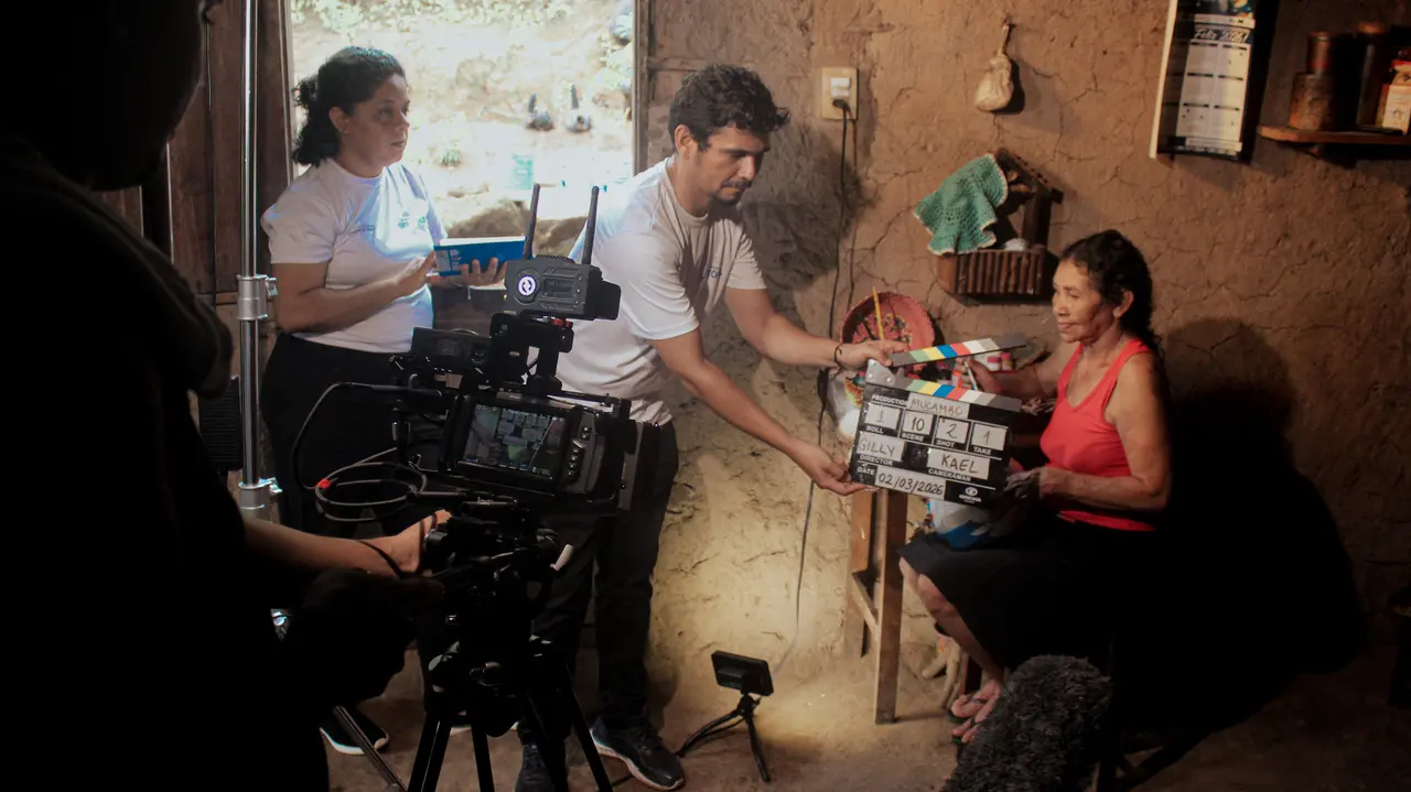 A equipe de filmagem prepara a gravação de uma cena no interior de uma casa com paredes de taipa, destacando um homem que estende a claquete em direção a uma senhora sentada. Em primeiro plano, a câmera de cinema profissional posicionada no tripé captura o enquadramento, enquanto o restante da produção acompanha os ajustes sob uma iluminação direcionada.