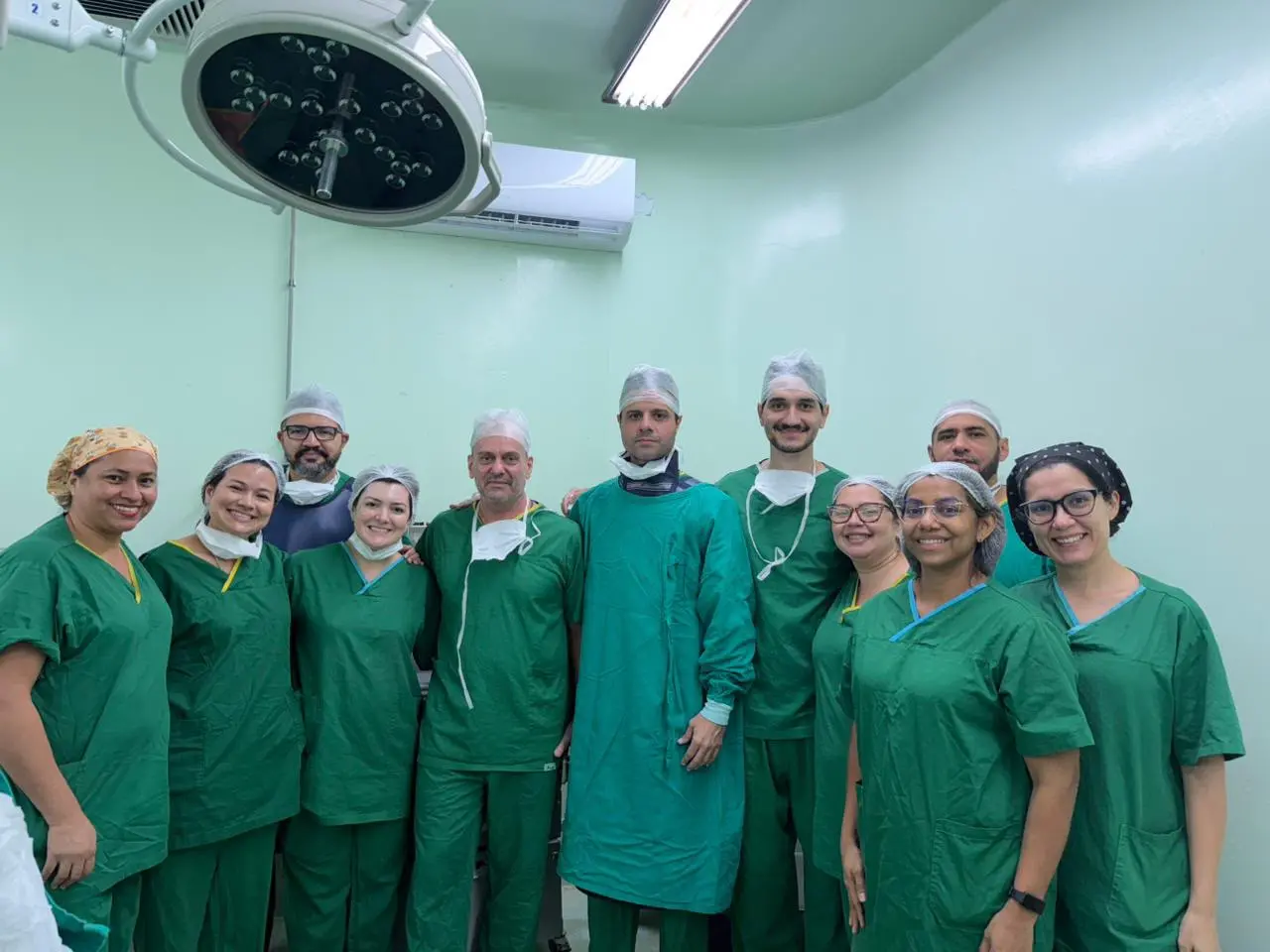 Fotografia colorida em plano médio de uma equipe médica composta por dez profissionais, entre homens e mulheres, posando sorridentes lado a lado dentro de uma sala de cirurgia. Todos vestem uniformes de privativo cirúrgico em tons de verde-escuro, toucas e máscaras descartáveis abaixadas no pescoço. O ambiente possui paredes de tom verde-claro, um aparelho de ar-condicionado ao fundo e uma grande luminária cirúrgica de teto visível no canto superior esquerdo.