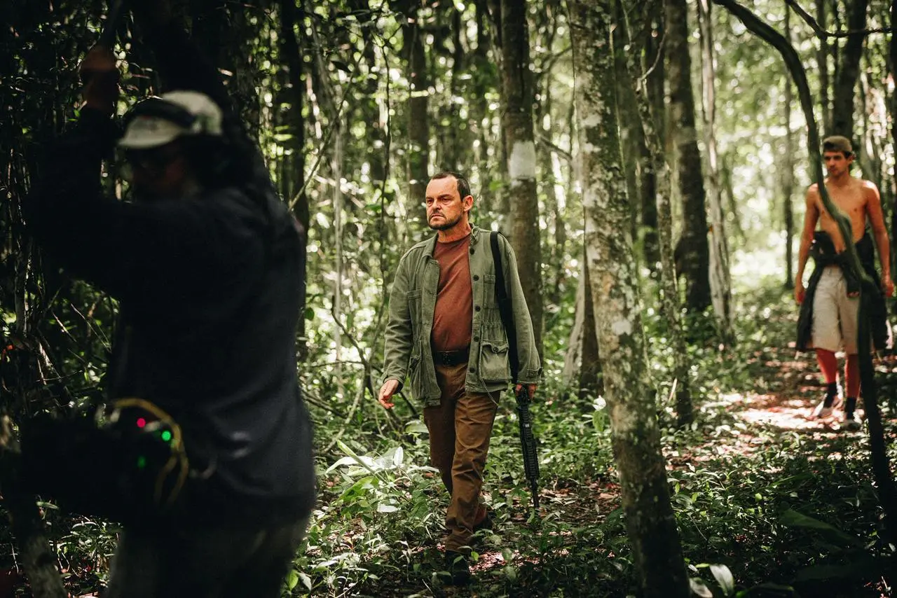 O ator Matheus Nachtergaele caminha por uma trilha na floresta densa segurando uma arma longa, vestindo calça marrom, camiseta no mesmo tom e jaqueta verde-oliva. Ele é acompanhado ao fundo por um jovem sem camisa, enquanto um integrante da equipe de filmagem de costas aparece desfocado em primeiro plano à esquerda.