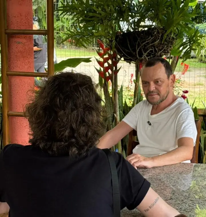 O ator Matheus Nachtergaele, vestindo camiseta branca, está sentado à mesa conversando com uma pessoa de costas com cabelos cacheados e camiseta preta. Ao fundo, uma varanda decorada com plantas tropicais e folhagens verdes emoldura o ambiente externo iluminado.