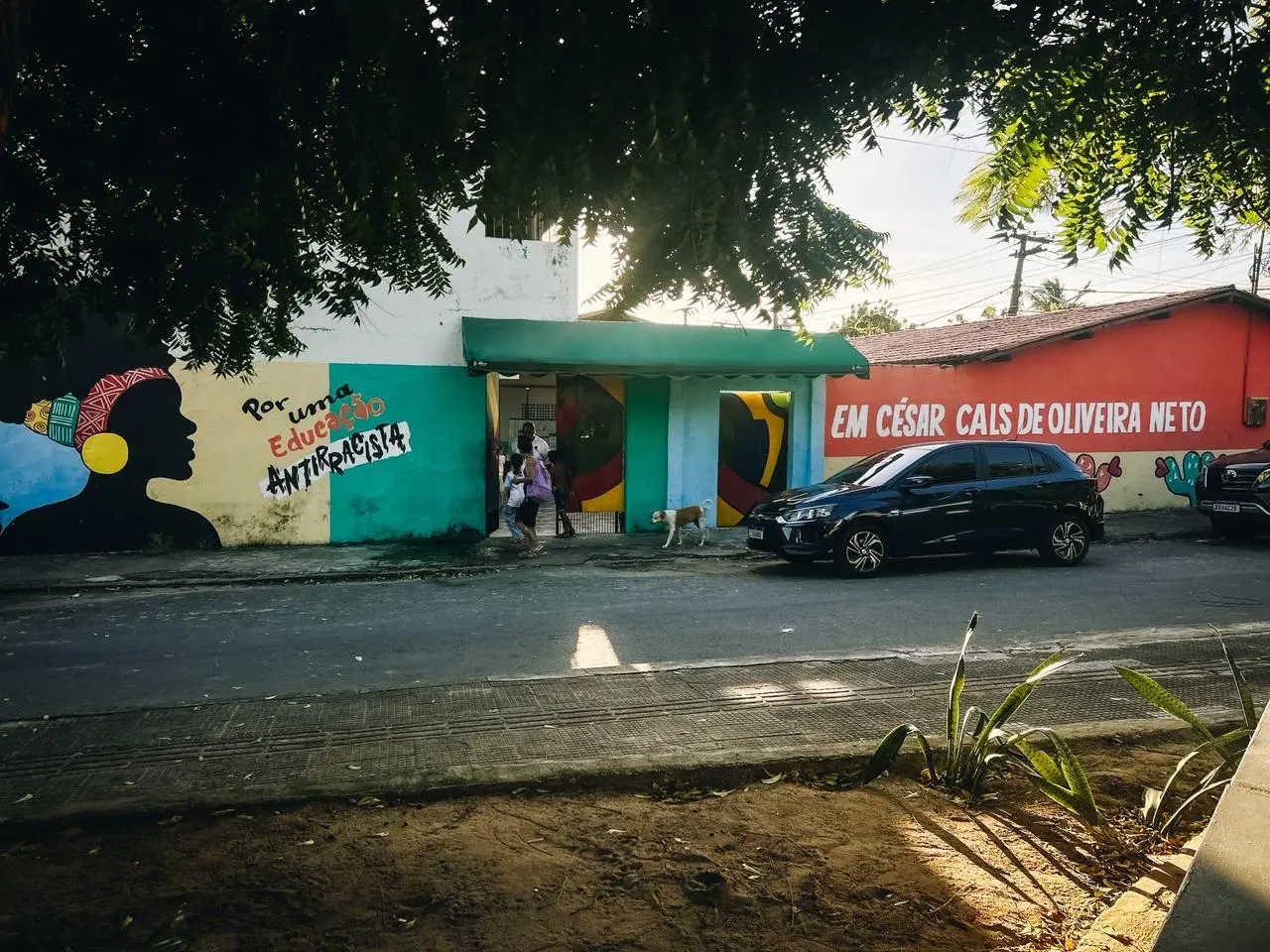 Fachada do CEI César Cals, que tem paredes pintadas de azul, vede e laranja. Do lado do portão da escola, que está aberto, uma pintura de uma mulher negra de turbante com os dizeres 