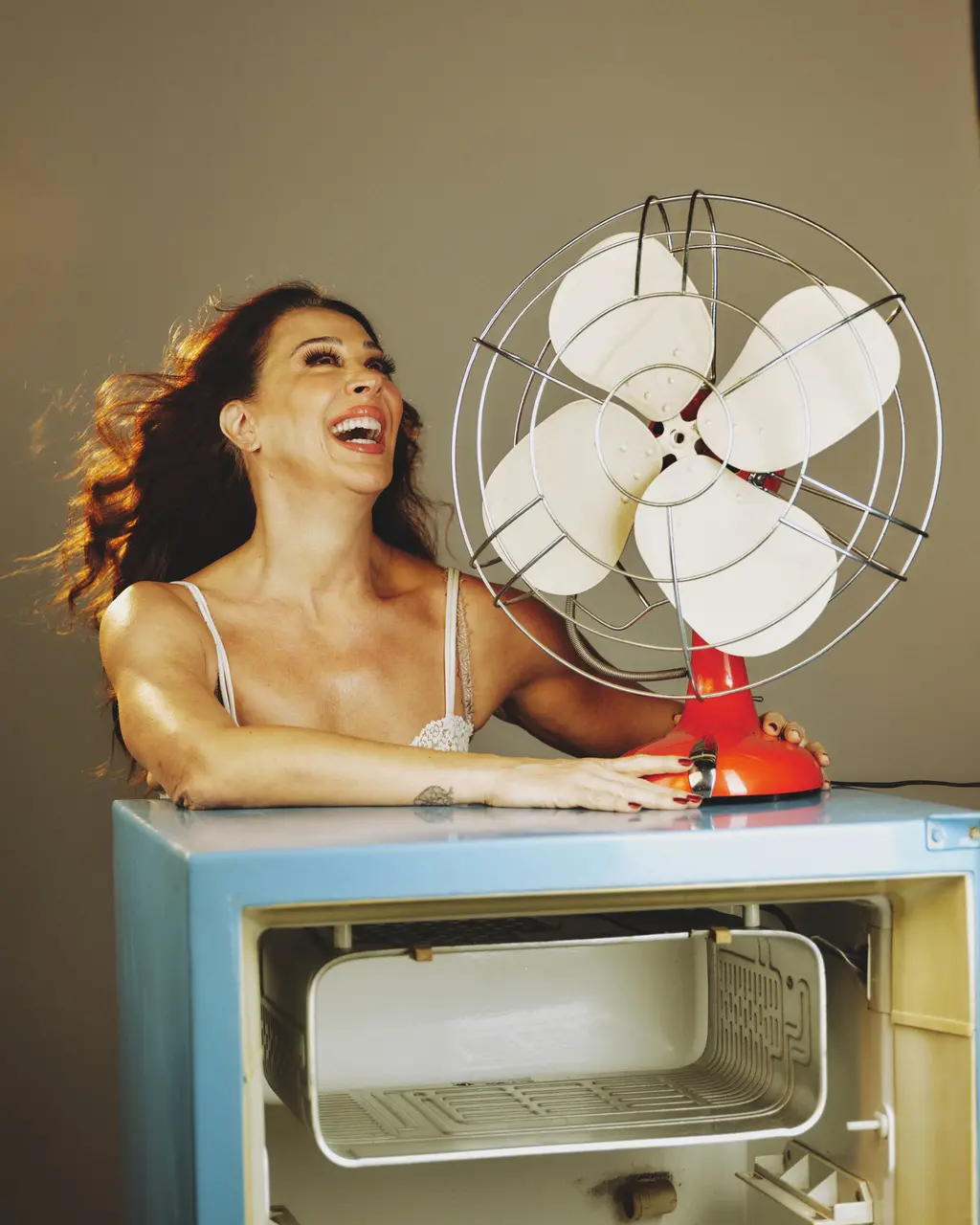 A atriz Claudia Raia aparece sorrindo e com a cabeça inclinada para trás enquanto se apoia em cima de uma geladeira azul vintage com a porta aberta. Seus cabelos estão ao vento, direcionados por um grande ventilador de mesa antigo com base vermelha que ela segura com as duas mãos, compondo um cenário divertido sobre um fundo neutro.