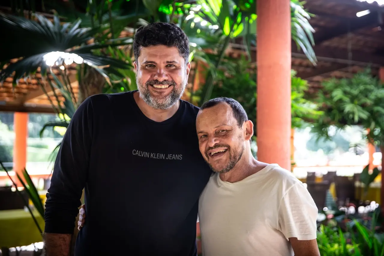 O diretor Petrus Cariry e o ator Matheus Nachtergaele sorriem abraçados em um ambiente coberto ao ar livre, cercados por folhagens verdes e pilares de tom terracota. Petrus veste uma camiseta de manga longa preta e o ator usa uma camiseta branca, registrando um momento de descontração e parceria nos bastidores do filme.