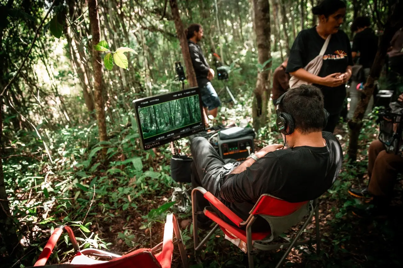 O diretor Petrus Cariry aparece de costas, sentado em uma cadeira de praia vermelha e usando fones de ouvido enquanto monitora uma cena da floresta em uma tela portátil montada no set. Ao redor dele, a vegetação densa envolve outros membros da equipe técnica que circulam e trabalham nos bastidores da gravação.