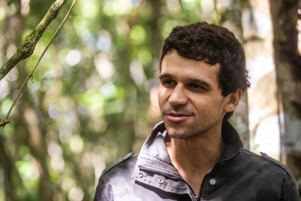 O ator cearense Lucas Galvino, com cabelos curtos e leve barba rala, sorri sutilmente de perfil, vestindo uma jaqueta de gola alta em tom escuro. O plano médio destaca sua expressão tranquila contra um fundo de floresta bastante desfocado, onde a luz do sol se filtra entre as folhas criando pontos luminosos.