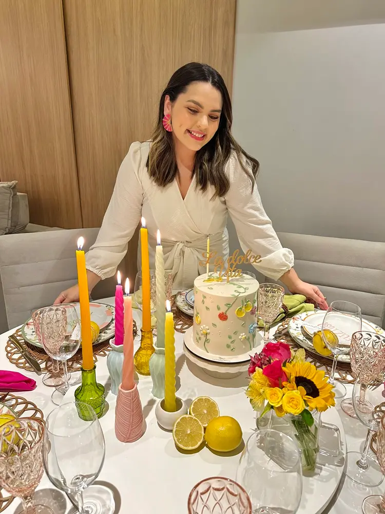 Na imagem, uma mulher jovem, de pele clara e cabelos castanhos na altura dos ombros, está de pé atrás de uma mesa de jantar redonda e elegantemente decorada. Ela veste uma blusa ou vestido transpassado na cor off-white, com mangas longas levemente bufantes, e usa brincos grandes em formato de leque na cor rosa vibrante. Ela olha para baixo com um sorriso suave, contemplando a mesa. Sobre a mesa branca, há um bolo de aniversário cilíndrico e alto, decorado com flores delicadas, ramos verdes pintados e um topo de bolo dourado escrito 