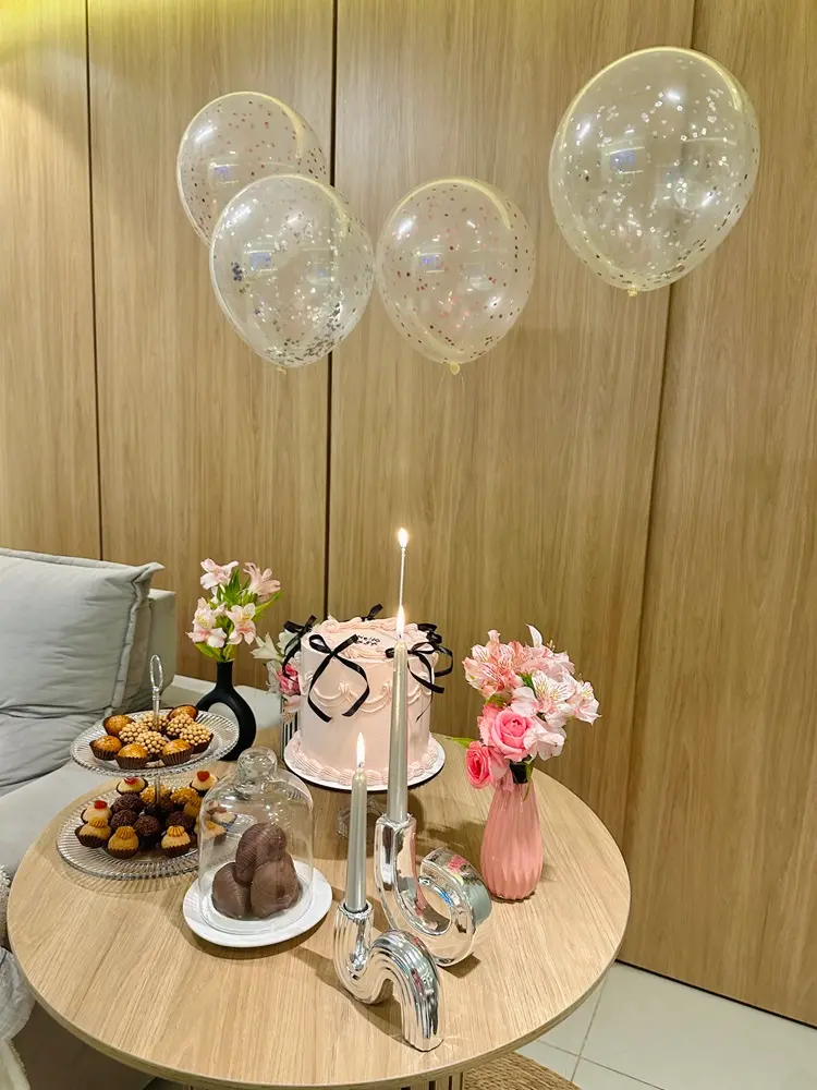 Na imagem, uma mesa redonda de madeira clara decorada para uma festa de aniversário, posicionada em frente a uma parede de painel de madeira. No centro da mesa, há um bolo cilíndrico rosa claro decorado com pequenos laços pretos de fita e detalhes em glacê. À frente do bolo, destaca-se um castiçal de metal prateado com um design moderno e ondulado, segurando duas velas finas e prateadas acesas. À esquerda do bolo, há uma torre de vidro de dois andares com docinhos de festa (como brigadeiros gourmets) e, mais à frente, uma cúpula de vidro transparente cobrindo ovos de chocolate. A decoração inclui dois vasos de flores: um vaso rosa canelado com rosas e astromélias cor-de-rosa à direita, e um vaso preto fosco menor com flores claras à esquerda. Flutuando acima da mesa, quatro balões transparentes cheios de confetes prateados e rosados completam o cenário festivo. À esquerda, nota-se o canto de um sofá cinza claro.