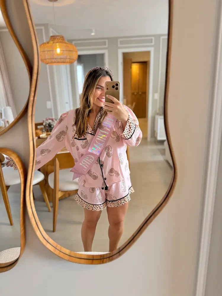 Na imagem, uma selfie no espelho de corpo inteiro tirada por uma mulher jovem de pele clara e cabelos castanhos claros. Ela está sorrindo alegremente, posicionada no centro do reflexo de um espelho com moldura de madeira de linhas sinuosas e orgânicas. Ela veste um pijama curto de botões na cor rosa claro com estampa de leopardo e detalhes pretos nas bordas. Atravessada em seu corpo, há uma faixa rosa com glitter escrita 