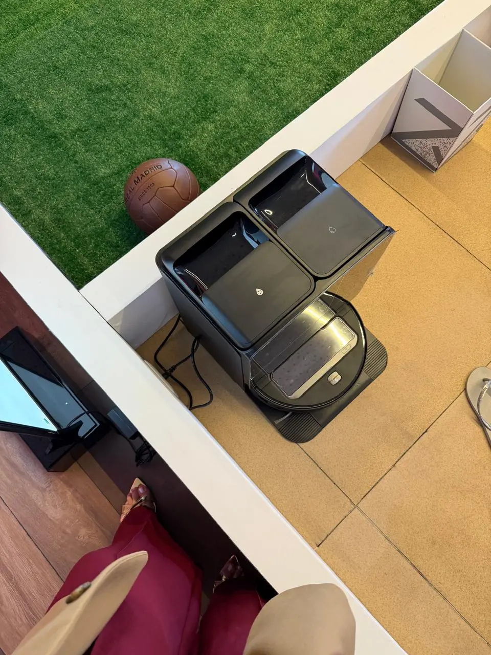 Na imagem, foto tirada de um ângulo superior (vista aérea) mostrando um robô aspirador de pó e água preto acoplado à sua estação de limpeza retangular inteligente, também preta. O aparelho está posicionado sobre um piso de placas bege claro. No canto esquerdo superior, separado por uma mureta branca baixa, há uma área com gramado sintético verde onde aparece uma bola de futebol de couro marrom de estilo retrô com a inscrição 