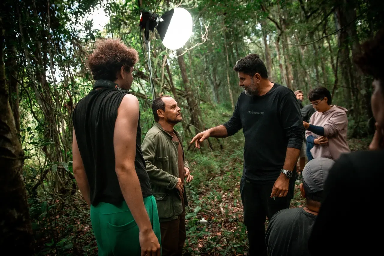 O diretor Petrus Cariry, à direita, gesticula enquanto orienta o ator Matheus Nachtergale, de casaco verde, no centro da imagem. À esquerda, um jovem de camiseta regata preta compõe a cena no meio de uma floresta densa. O registro de bastidores destaca um grande refletor de luz esférico e aceso ao fundo, iluminando a equipe técnica que acompanha a dinâmica de ensaio sobre o chão de folhas secas.