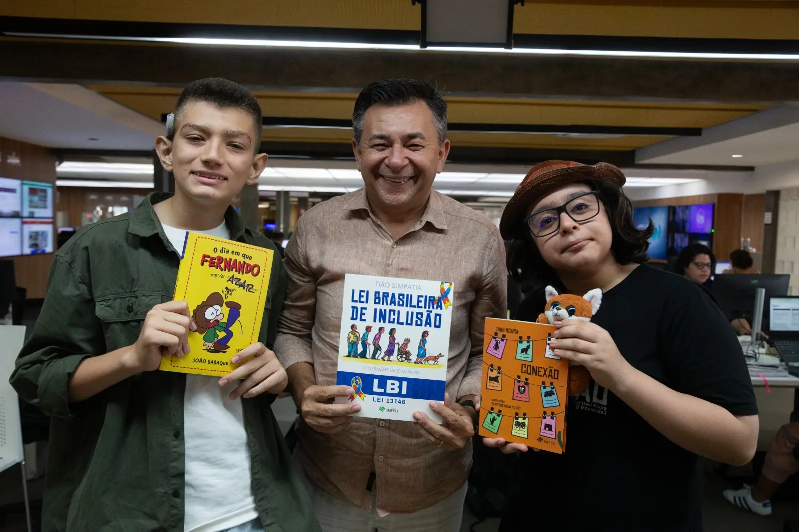 Na imagem, três pessoas de pele clara estão de pé, lado a lado, sorrindo para a câmera em um ambiente de escritório e segurando livros. À esquerda, um jovem de cabelos curtos raspados nas laterais veste uma camisa de botão verde-oliva aberta sobre uma camiseta branca; ele segura um livro amarelo intitulado 