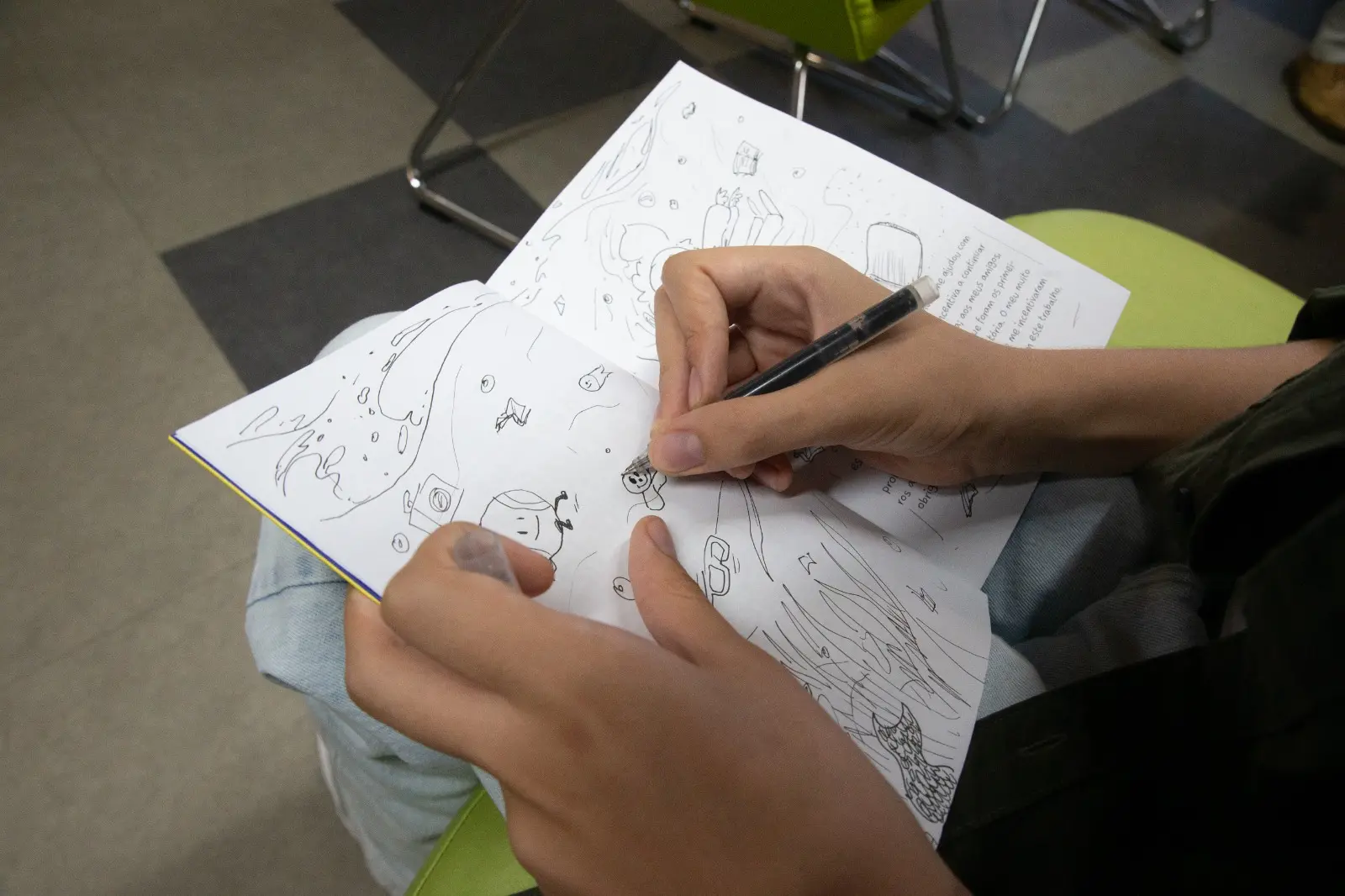 Na imagem, foto em plano detalhe e de ângulo superior mostrando as mãos de uma pessoa desenhando em um pequeno livro ou caderno aberto sobre o próprio colo. Com a mão direita, ela segura uma caneta esferográfica de corpo transparente e ponta fina, adicionando traços a uma ilustração em preto e branco cheia de pequenos personagens de traços simples, ondas e elementos lúdicos. A mão esquerda apoia a base do livro, segurando a tampa transparente da caneta entre os dedos. A pessoa veste uma calça jeans clara e uma camisa verde-oliva. O livro está aberto em uma página que combina esses desenhos feitos à mão com um bloco de texto impresso ao fundo. A cena se passa em um ambiente interno com piso cinza e detalhes de cadeiras com assento verde.